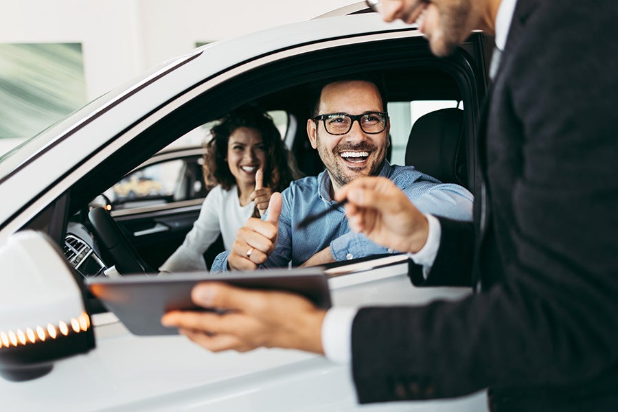 a man and woman sitting in a vehicle at a dealership with a sales person standing next to them